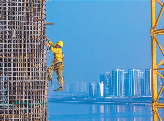 奮戰(zhàn)在百米索塔之上的建設(shè)者身影，與遠(yuǎn)處的高樓大廈遙呼相應(yīng)。